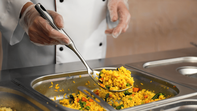 Midsection of chef body in white coat with black buttons, wearing clear gloves, serving yellow rice with vegetables out of serving well