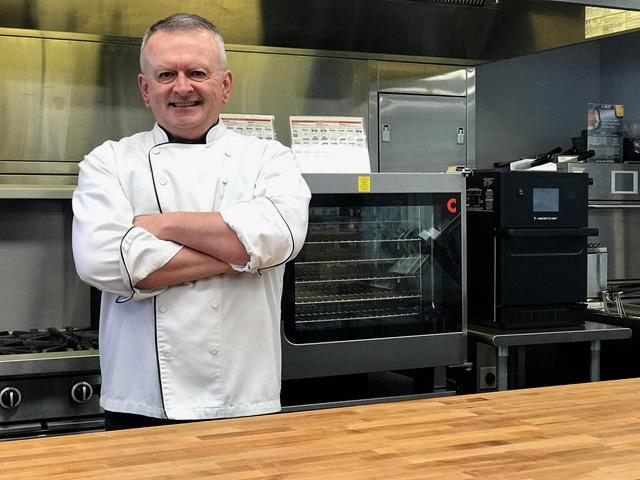Chef Doug, in a white jacket with arms crossed, stands in front of kitchen equipment Chef Doug, in a white jacket with arms crossed, stands in front of kitchen equipment